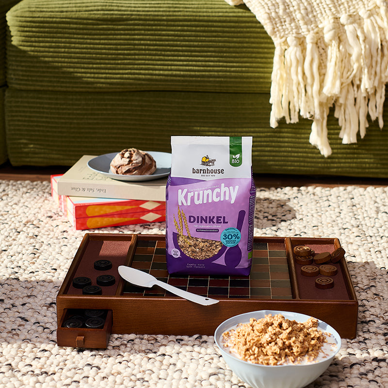 Lila Beutel Krunchy Dinkel weniger Zucker von Barnhouse, der gemeinsam mit einem weißen Löffel auf einem braun-schwarzen Dame-Spielbrett steht. Im Vordergrund steht eine weiße Schüssel mit Milch und dem besagten Bio-Knuspermüsli. Im Hintergrund sind noch eine grüne Couch mit weißer Strickdecke, ein weiß-schwarzer Teppich sowie ein Turm aus einem zusammengeklappten Backgammon-Spielbrett und einem Buch zu sehen, auf dem ein blauer Teller mit einem kleinen Brotlaib steht.