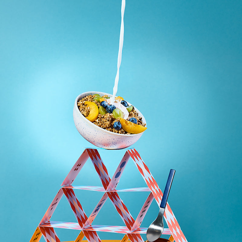 Kartenhaus aus roten Spielkarten, auf dem eine Schüssel mit Krunchy Dinkel weniger Zucker von Barnhouse, Trauben, Weintrauben und Pfirsich steht. Von oben fließt ein Milchstrahl in das Bio-Knuspermüsli. Rechts ist noch ein blau-silberner Löffel zu sehen.