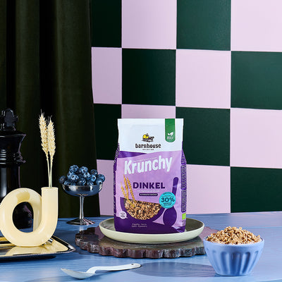 Lila Beutel Krunchy Dinkel weniger Zucker von Barnhouse in grünlichem Teller auf geriffelter Marmorplatte. Rechts vorne steht eine blaue Schüssel mit Milch und besagtem Bio-Knuspermüsli. Außerdem stehen noch ein silbernes Aperitif-Glas, ein weißer Löffel, eine gelbe, geschwungene Vase mit zwei Dinkelhalmen auf einem silbernen Tablett sowie eine schwarze Pfeffermühle in Form eines Schach-Königs auf blauem Holztisch. Im Hintergrund eine Wand mit dunkelgrün-gräulichem Schachmuster sowie dunkelgrünem Vorhang.