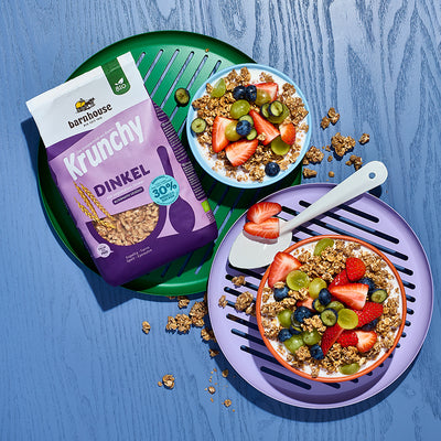 Blick von oben auf einen lila Beutel Krunchy Dinkel weniger Zucker von Barnhouse, der in einem grünen Teller liegt. Daneben eine blaue Müslischüssel mit diesem Bio-Knuspermüsli, Milch, Trauben, Heidelbeeren und Erdbeeren. Daneben ein lila Teller mit einer roten Schüssel, die mit dem gleichen Inhalt gefüllt ist. Daneben liegt ein weißer Löffel. Auf dem blauen Holztisch liegen noch ein paar Cluster vom Dinkel-Krunchy. 