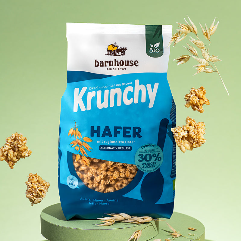 Blauer Beutel Krunchy Hafer weniger Zucker von Barnhouse auf einem grünen, runden Sockel. Außenrum fliegen Cluster dieses Bio-Knuspermüslis und Haferhalme.