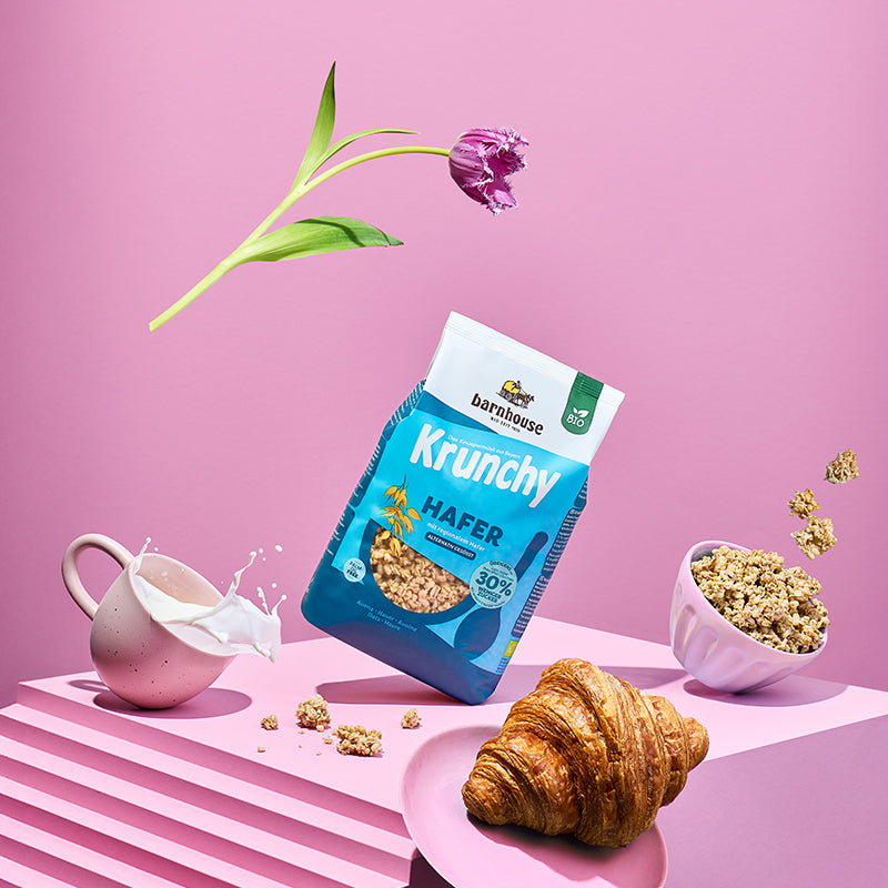 Blick voller Schwung von vorne auf ein rosa Treppe. Alle Elemente auf dem Bild "fliegen" durch die Luft: Blau-weißer Beutel Krunchy Hafer weniger Zucker von Barnhouse, fliederfarbene Schale mit diesem Bio-Knuspermüsli, Krunchy-Cluster, cremefarbene Tasse mit Milch, rosa Teller mit Croissant sowie eine Tulpe mit lila Blütenknospe.