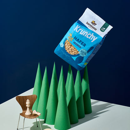 10 dunkelgrüne, hohe Kegel. Auf einem der Kegel steckt ein blauer Beutel Krunchy Hafer weniger Zucker von Bio-Knuspermüsli Hersteller Barnhouse. Vorne links ist noch eine Miniatur-Holzstuhl mit zwei weißen Schachfiguren: König und Bauer.
