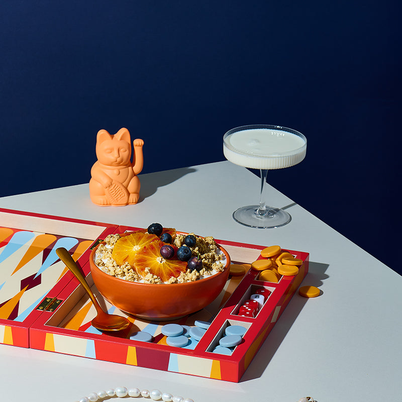 Buntes Backgammon-Spielbrett, in dem neben den farbigen Spielfiguren und Würfeln auch ein bronzener Löffel sowie eine orange Schüssel mit Milch, Krunchy Hafer weniger Zucker von Bio-Knuspermüsli Hersteller Barnhouse sowie Heidelbeeren und Blutorangen-Scheiben steht. Im Hintergrund sind noch eine winkende Katze (Maneki-neko) und ein Martini-Glas mit Milch zu sehen.
Vorne blitzt eine weiße Halskette ins Bild.