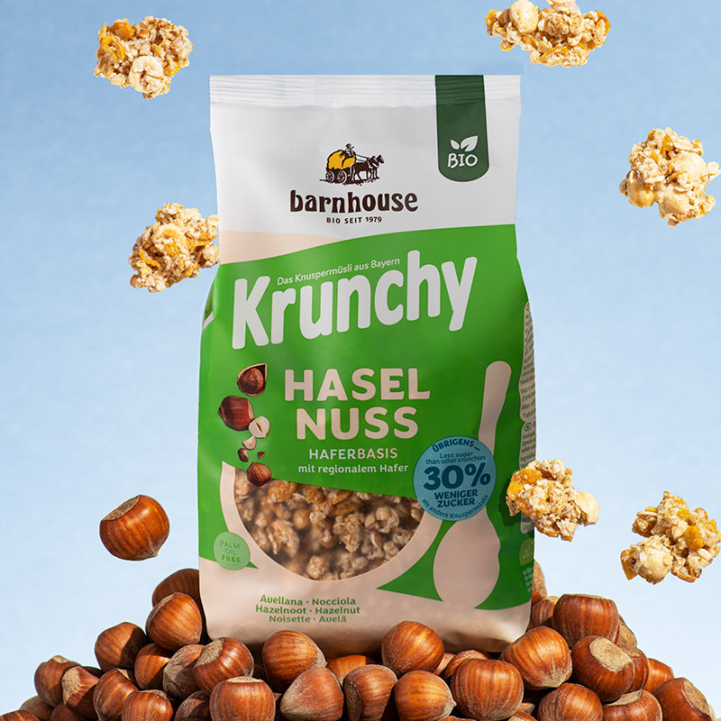 Grün-beiger Beutel Krunchy Haselnuss weniger Zucker von Bio-Knuspermüsli-Hersteller Barnhouse auf einem Berg aus Haselnüssen. Außenrum fliegen Cluster von diesem Krunchy.