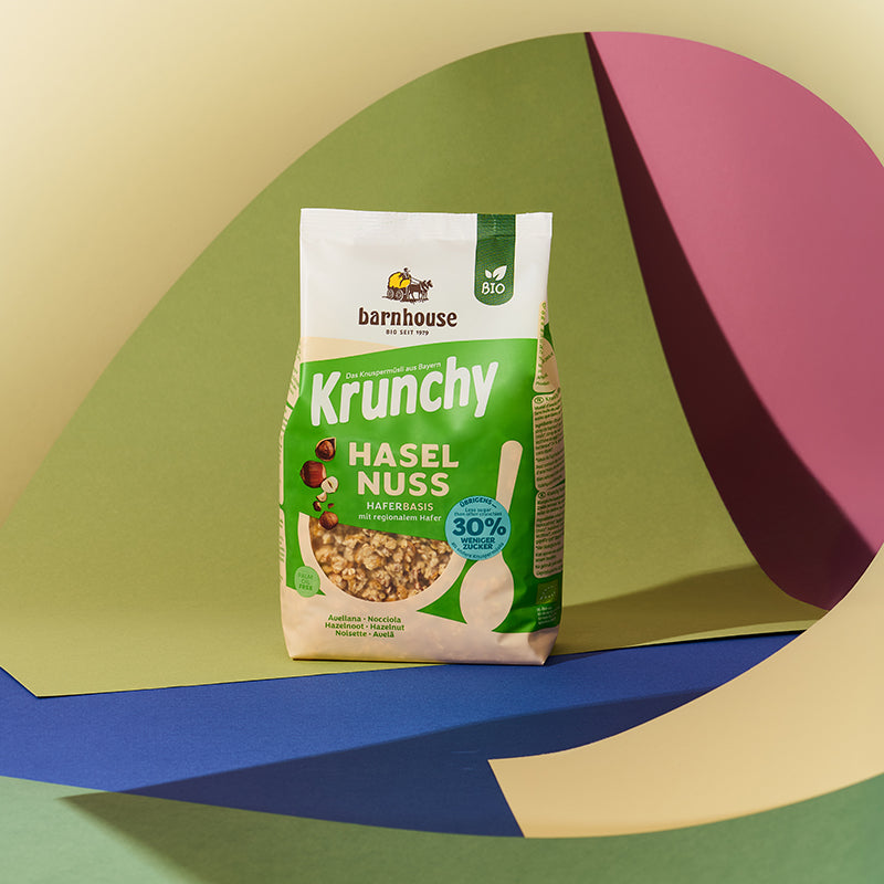 Grün-beiger Beutel Krunchy Haselnuss weniger Zucker von Bio-Knuspermüsli Hersteller Barnhouse vor einem bunten Hintergrund mit einer gelben, grünen, roten, blauen und brauen Fläche.