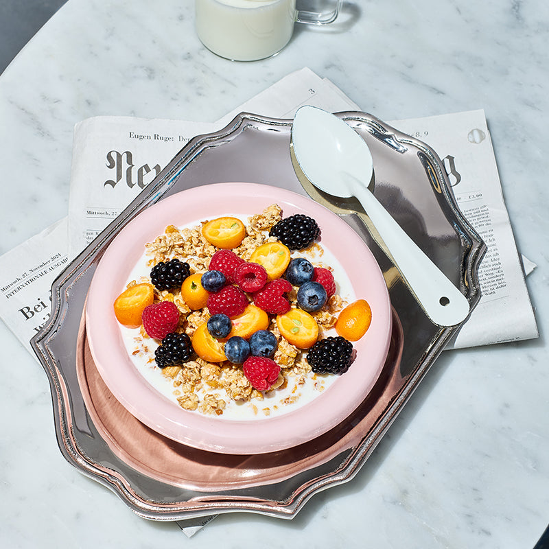 Silbernes Tablett, auf dem eine rosa Schüssel mit Milch, Krunchy Haselnuss weniger Zucker, Himbeeren, Brombeeren, Heidelbeeren und Physalis steht. Daneben ein weißer Löffel.  Das Tablett liegt auf einer Zeitung, die auf einem Marmortisch liegt. Am oberen Bildrand ist noch ein Teil eines durchsichtigen Glases mit Milch zu sehen.