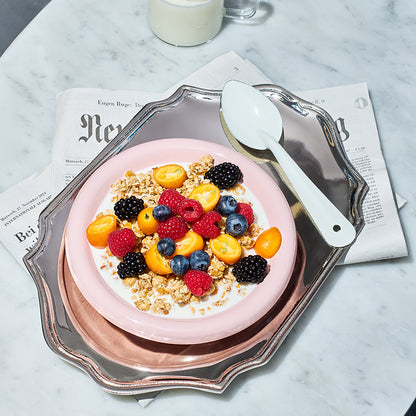 Silbernes Tablett, auf dem eine rosa Schüssel mit Milch, Krunchy Haselnuss weniger Zucker, Himbeeren, Brombeeren, Heidelbeeren und Physalis steht. Daneben ein weißer Löffel.  Das Tablett liegt auf einer Zeitung, die auf einem Marmortisch liegt. Am oberen Bildrand ist noch ein Teil eines durchsichtigen Glases mit Milch zu sehen.