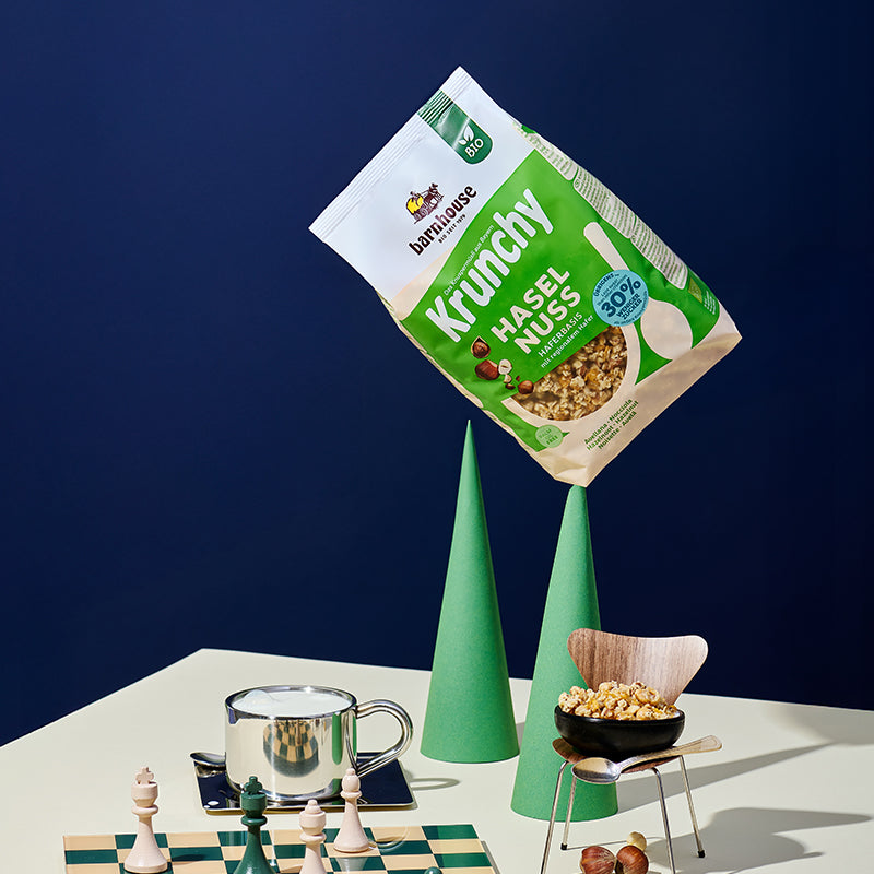 Weißer Holztisch, auf dem zwei grüne, hohe Kegel stehen. Auf dem einen ist ein grün-beiger Beutel Krunchy Haselnuss weniger Zucker von Barnhouse aufgespießt. Daneben stehen eine Espressotasse mit Milch, ein Löffel, ein grün-beiger Schachbrett mit einer Paar Figuren sowie auf Miniatur-Holztisch, auf dem eine schwarze Schüssel mit dem Nuss-Müsli steht. Außerdem liegt noch ein silberner Löffel auf dem Stuhl. Auf dem Tisch liegen noch Haselnüsse.