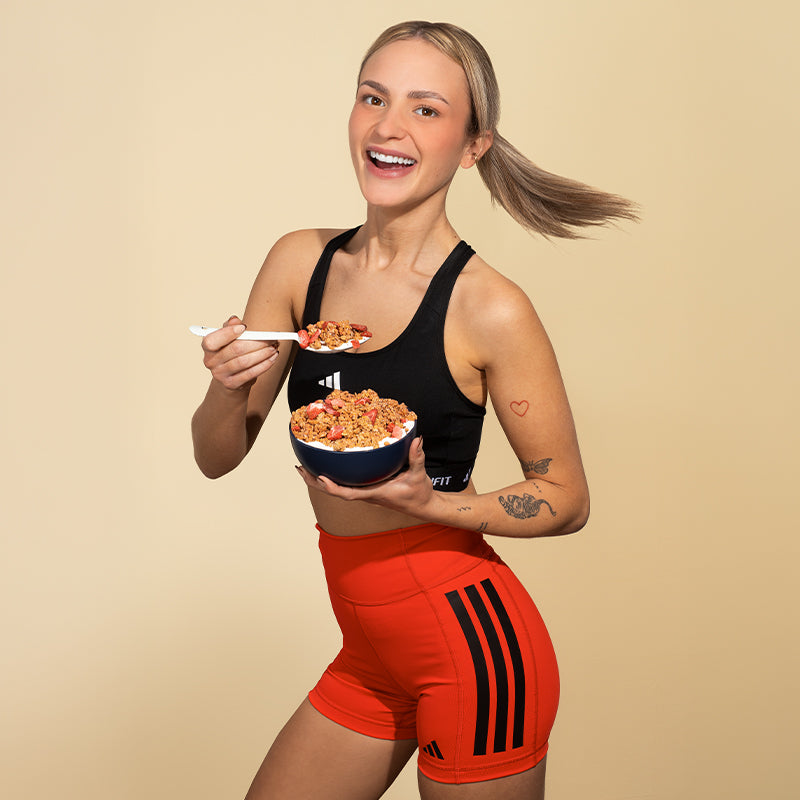 Eine junge, blonde und tätowierte Frau im Sport-Outfit, die in die Kamera strahlt. In der linken Hand hält sie eine schwarze Schüssel mit Krunchy High Protein von Barnhouse mit Milch und in der rechten Hand einen weißen Löffel, der mit diesem Bio-Knuspermüsli gefüllt ist.
