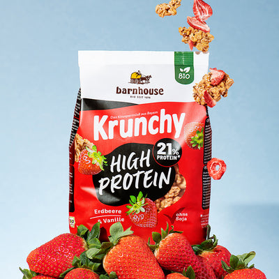 Beutel Krunchy High Protein von Bio-Knuspermüsli-Hersteller Barnhouse, der auf einem Berg aus Erdbeeren steht. Von oben fallen Knuspermüsli-Cluster und Erdbeerscheiben nach unten.