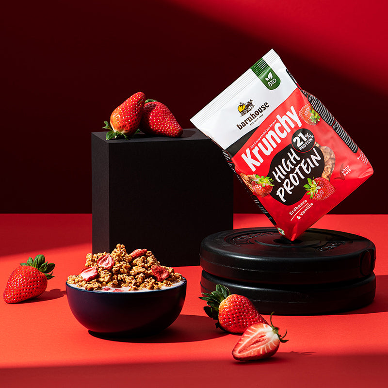 Beutel Krunchy High Protein von Barnhouse, der im 45 Grad Winkel (nach links geneigt) auf 2 Hantelgewichten steht. Links daneben befindet sich ein schwarzes Achteck, auf dem 2 Erdbeeren liegen. Auf dem roten Untergrund befinden sich noch 2 weitere, ganze Erdbeeren sowie ein Erdbeer-Hälfte. Links vorne steht eine dunkle Schale mit dem besagten Bio-Knuspermüsli (Cluster + Erdbeerscheiben) und Milch.