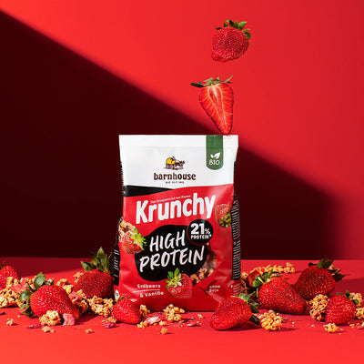 Blick von vorne auf einen Beutel Krunchy High Protein von Barnhouse, der auf auf einer roten Fläche steht. Von oben fallen 2 Erdbeeren (eine ganze und eine Hälfte) herunter und außenrum liegen jede Menge Erdbeeren sowie Cluster vom erwähnten Bio-Knuspermüsli.