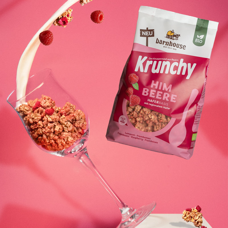Beutel Krunchy Himbeere, der rechts neben einem Weinglas schwebt, das links von einem weißen Sockel fällt. Das Weinglas ist mit Clustern des Himbeer-Müslis befüllt. Vom oberen Bildrand fließt ein Milchstrahl ins Glas und weitere Cluster sowie Himbeeren fliegen herum.