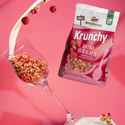 Beutel Krunchy Himbeere, der rechts neben einem Weinglas schwebt, das links von einem weißen Sockel fällt. Das Weinglas ist mit Clustern des Himbeer-Müslis befüllt. Vom oberen Bildrand fließt ein Milchstrahl ins Glas und weitere Cluster sowie Himbeeren fliegen herum.