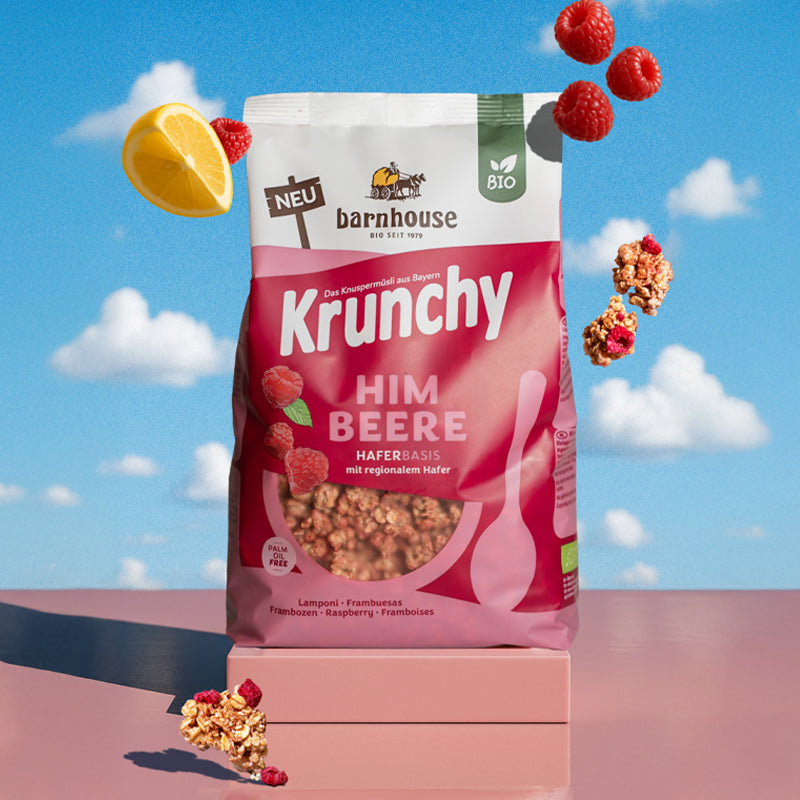 Beerenfarbiger, rosa-weißer Beutel Krunchy Himbeere von Bio-Knuspermüsli-Hersteller Barnhouse auf einem eckigen Sockel. Von oben fallen Knuspermüsli-Cluster, Himbeeren &amp; 1 Zitronen-Viertel vor einem Hintergrund mit blauem Himmel und Wolken nach unten.