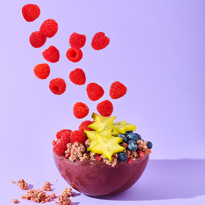 Beerenfarbene Schüssel vor lila Hintergrund, die mit Krunchy Himbeere, Heidelbeeren, Sternenfrucht und Himbeeren befüllt ist. Von oben fallen weitere Himbeeren in die Schüssel und links daneben liegen Krunchy Cluster.