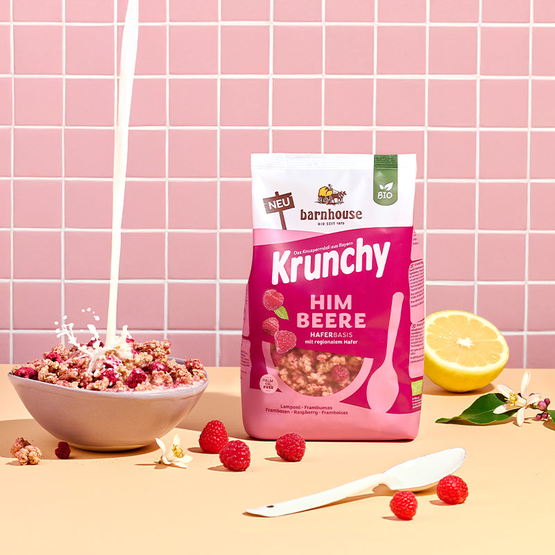 Blick von vorne auf den beerenfarbigen, rosa-weißer Beutel Krunchy Himbeere von Bio-Knuspermüsli-Hersteller Barnhouse, der auf einem beigen Tisch steht. Links daneben steht eine Schüssel mit diesem Müsli, in die von oben ein Milchstrahl fließt. Daneben liegen eine halbe Zitrone, Himbeeren, Krunchy-Cluster sowie ein weißer Löffel und zwei weiße Blüten. Im Hintergrund ist eine rosa geflieste Wand zu sehen.