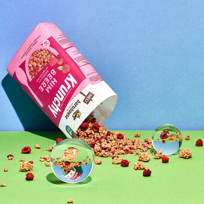Blick von vorne auf den beerenfarbigen, rosa-weißen Beutel Krunchy Himbeere von Bio-Knuspermüsli-Hersteller Barnhouse. Der Beutel ist auf den Kopf gedreht und geöffnet, wodurch jede Menge Krunchy-Cluster und Himbeeren herausrieseln und auf den grünen Untergrund fallen. Zwischendrin liegen 2 Glaskugeln, die Elemente des Bildausschnittes spiegeln. Die Wand im Hintergrund ist blau.