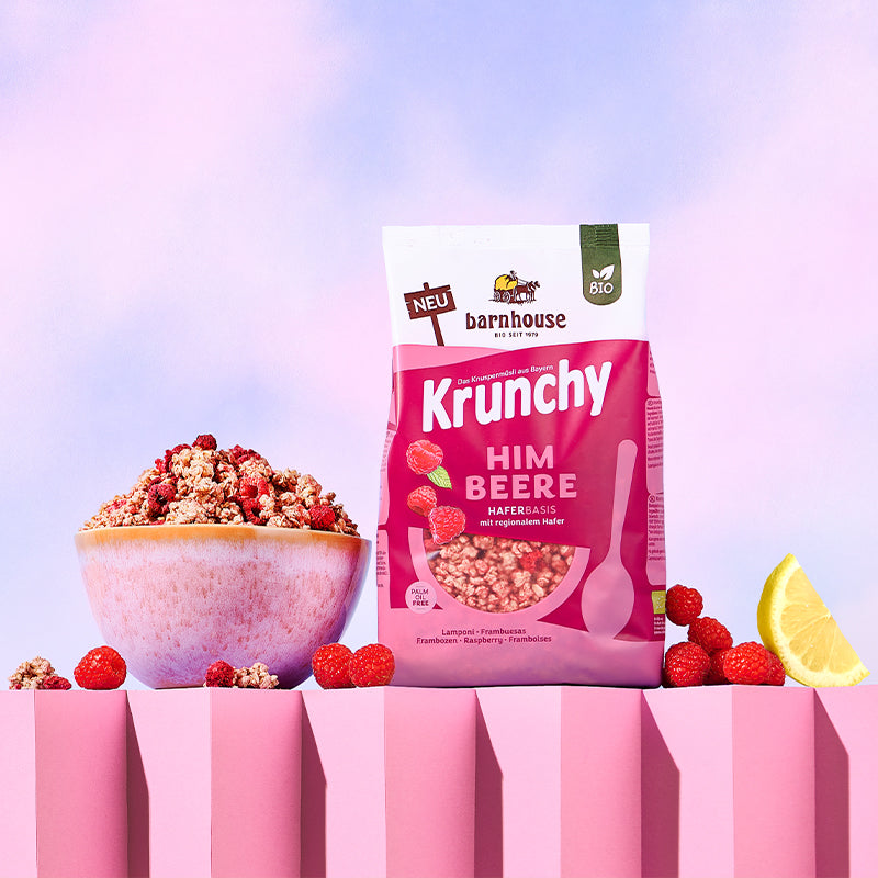 Blick von vorne auf den beerenfarbigen, rosa-weißer Beutel Krunchy Himbeere von Bio-Knuspermüsli-Hersteller Barnhouse, der auf einem gezackten, rosafarbenen Sockel steht. Links daneben steht eine Schüssel mit diesem Müsli, die von Himbeeren und Krunchy-Clustern umgeben ist. Rechts daneben liegen ein paar Himbeeren sowie ein Achtel einer Zitrone. Der Hintergrund ist hell.