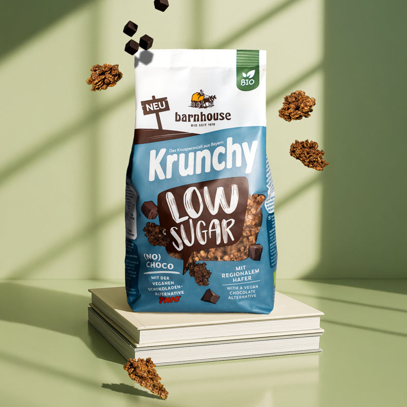 Blau-braun-weißer Beutel Krunchy Low Suagr (No) Choco von Bio-Knuspermüsli-Hersteller Barnhouse, der auf 2 hellen Büchern steht. Von oben fallen Knuspermüsli-Cluster und THIC-Schoko-Stücke vor einem grünen Hintergrund nach unten. Von außen strahlt Sonne durch ein Fenster in den Raum.