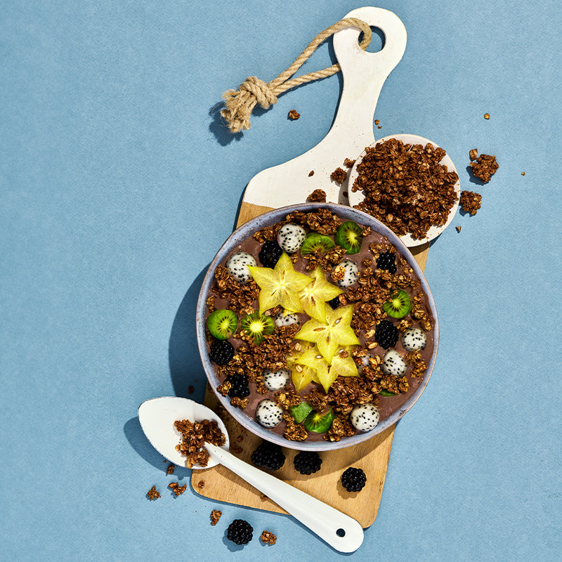 Blick von oben auf ein weiß-braunes Holzbrett, auf dem eine Schüssel steht, die mit Krunchy Low Sugar (No) Choco von Bio-Knuspermüsli-Hersteller Barnhouse, Sternenfrucht, Brombeeren, Kiwi und Drachenfrucht befüllt ist. Daneben steht eine weiße Schüssel mit diesem Krunchy sowie ein weißer Löffel, auf dem ebenfalls Cluster liegen. Auf dem hellblauen Hintergrund liegen weitere Cluster sowie Brombeeren verteilt.