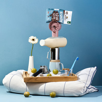 Blick von vorne auf ein Holzbrett, das auf einem blau-weiß karierten Kissen steht. Auf dem blauem Boden liegen 2 Birnen. Auf dem Holzbrett stehen ein Glas mit blauer Zahnbürste, eine weiße Vase mit weißer Blume und ein Kaffeesieb. Daneben ein Turm aus einem Buch, einer Tasse, einer Milchkanne, einer Milchflasche sowie einer Uhr und ganz oben liegt ein Beutel Krunchy Low Sugar (No) Choco von Bio-Knuspermüsli-Hersteller Barnhouse.