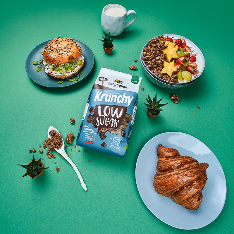 Blick von oben auf eine grüne Ablage, auf der ein blau-braun-weißer Beutel Krunchy Low Sugar (No) Choco von Bio-Knuspermüsli-Hersteller Barnhouse liegt. Außenrum stehen 3 Kakteen, ein Löffel mit Krunchy-Clustern sowie eine Schüssel mit diesem Müsli, Sternenfrucht, Weintrauben, Erdbeeren und Milch. Zudem stehen noch eine Tasse mit Milch, eine Teller mit Croissant sowie ein Teller mit einem Bagel auf dem Tisch.