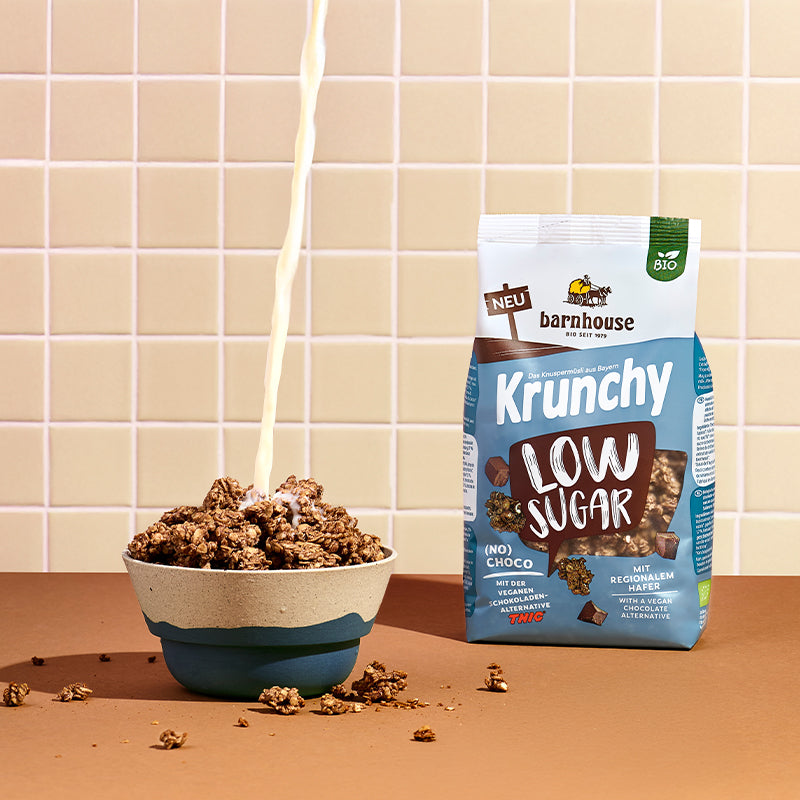 Blick von vorne auf einen blau-braun-weißen Beutel Krunchy Low Sugar (No) Choco von Bio-Knuspermüsli-Hersteller Barnhouse, der auf einer braunen Ablage steht. Links daneben eine blau-graue Schüssel mit diesem Krunchy, in die von oben ein Milchstrahl fließt. Außenrum liegen Krunchy-Cluster. Die Wand im Hintergrund ist weiß-beige gefliest.