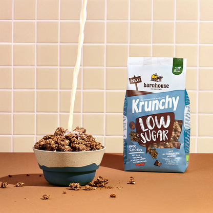 Blick von vorne auf einen blau-braun-weißen Beutel Krunchy Low Sugar (No) Choco von Bio-Knuspermüsli-Hersteller Barnhouse, der auf einer braunen Ablage steht. Links daneben eine blau-graue Schüssel mit diesem Krunchy, in die von oben ein Milchstrahl fließt. Außenrum liegen Krunchy-Cluster. Die Wand im Hintergrund ist weiß-beige gefliest.