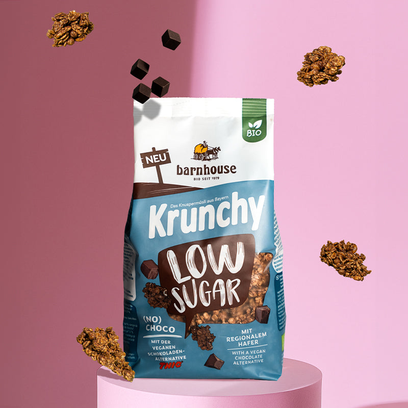 Blick von vorne auf einen blau-braun-weißen Beutel Krunchy Low Sugar (No) Choco von Bio-Knuspermüsli-Hersteller Barnhouse, der auf einer rosefarbenen Sockel steht. Außenrum schweben Krunchy-Cluster sowie Schokowürfel der veganen Schoko-Alternative THIC. Der Hintergrund ist ebenfalls rosa.