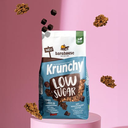 Blick von vorne auf einen blau-braun-weißen Beutel Krunchy Low Sugar (No) Choco von Bio-Knuspermüsli-Hersteller Barnhouse, der auf einer rosefarbenen Sockel steht. Außenrum schweben Krunchy-Cluster sowie Schokowürfel der veganen Schoko-Alternative THIC. Der Hintergrund ist ebenfalls rosa.