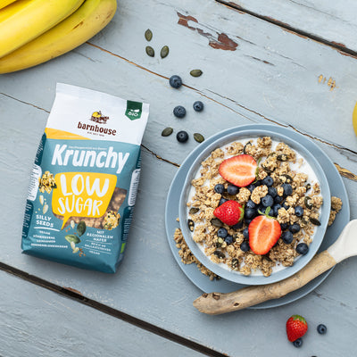 Blick von oben auf einen gelb-weiß-blauen Beutel Krunchy Low Sugar Silly Seeds von Bio-Knuspermüsli-Hersteller Barnhouse. Daneben steht eine Müslischale, die mit Milch und dem zuckerarmen Müsli befüllt ist. Frische Erdbeerscheiben und Heidelbeeren sorgen für eine schöne Farbkombi. Auf dem Unterteller liegt außerdem noch ein Holzlöffel sowie Müsli-Cluster. Auf dem blau gestrichenen Holztisch findet man noch Erdbeeren, Heidelbeeren, Bananen und Kürbiskerne verteilt.