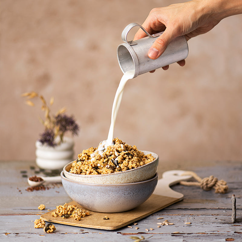 Blick auf zwei ineinander gestapelte Müslischalen. Die obere Schüssel ist mit Krunchy Low Sugar Silly Seeds von Bio-Knuspermüsli-Hersteller Barnhouse befüllt. Von oben schüttet eine Hand Milch aus einer kleinen Blechkanne über das Müsli. Die Schalen stehen auf einem dünnen Holzbrett, das auf einem blau gestrichenen Holztisch liegt. Auf dem Tisch sind noch Müsli-Cluster, Kürbiskerne, Leinsamen und Sonnenblumenkerne verteilt - die Zutaten des Müslis. Im Hintergrund steht  eine weiße Vase mit einem Haferhalm.