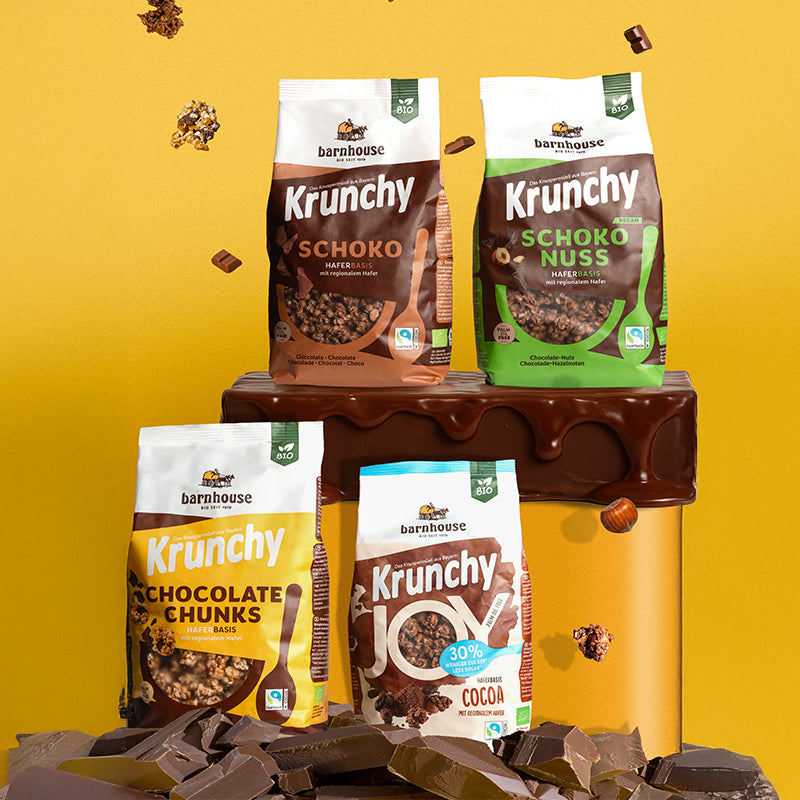 Blick von vorne auf die Beutel der vier Schoko-Krunchys Schoko-Nuss, Schoko, Chocolate Chunks und Joy Cocoa von Bio-Knuspermüsli-Hersteller Barnhouse. Krunchy Schoko und Schoko-Nuss stehen auf einem gleben, runden Sockel, der obenrum mit ordentlich Schokolade überzogen ist. Darunter stehen Krunchy Chocolate Chunks und Joy Cocoa auf Schoko-Stückchen. Von oben fallen Knuspermüsli-Cluster und Schoko-Stückchen vor gelbem Hintergrund nach unten.