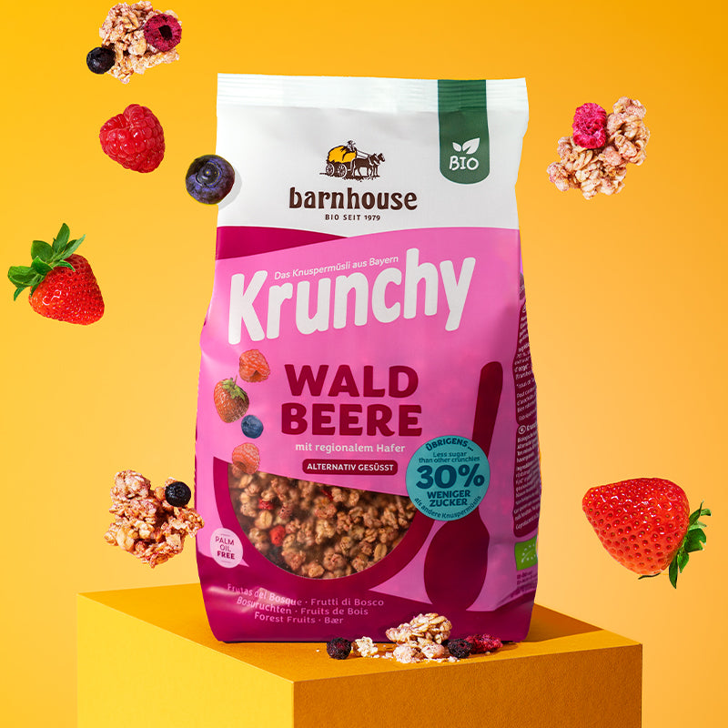 Rose-beerenfarbener Beutel Krunchy Waldbeere weniger Zucker von Barnhouse auf einem gelb-orangenen, quadratischen Sockel. Außenrum fliegen Cluster dieses Bio-Knuspermüslis sowie Heidelbeeren, Himbeere und Erdbeeren.