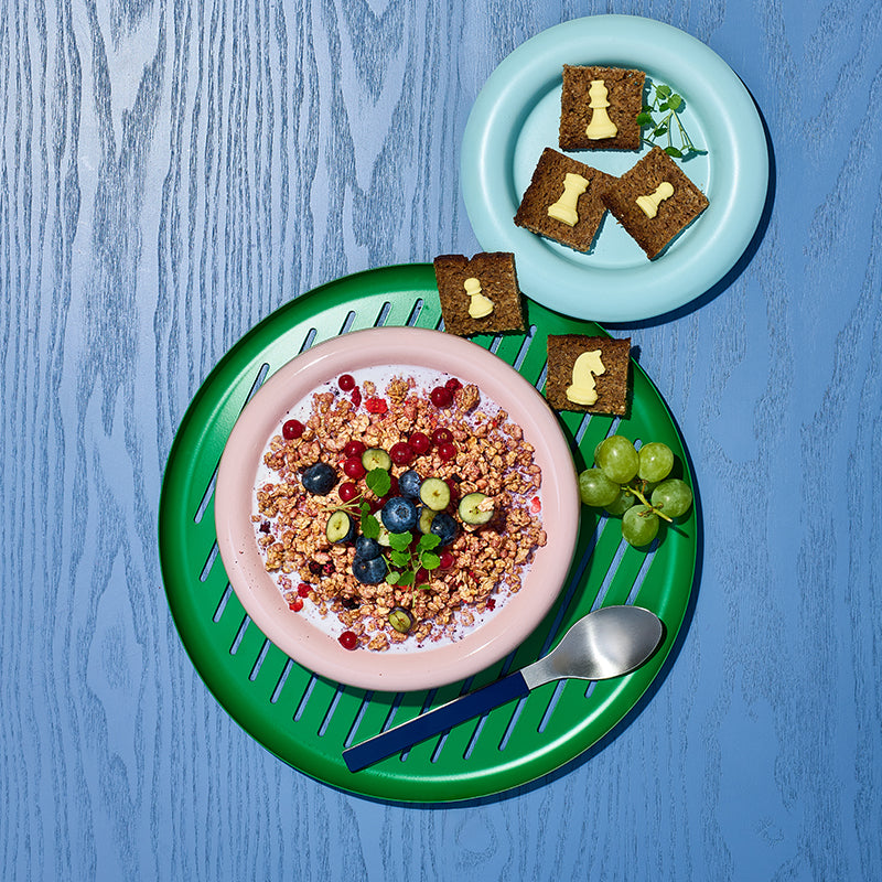 Blick von oben auf eine rosafarbene Müslischüssel mit Krunchy Waldbeere weniger Zucker von Bio-Knuspermüsli Hersteller Barnhouse, Johannisbeeren und Heidelbeeren. Die Schüssel steht auf einem grün-blauen Teller, auf dem auch noch ein blauer Löffel, grüne Weintrauben sowie kleine Vollkornbrot-Stücke mit Butter in Schachfiguren-Form liegen. Darüber liegt ein kleinerer, hellblauer Teller, auf dem weitere Vollkornbrot-Stücke mit Butter in Schachfiguren-Form liegen. Der Untergrund ist ein blauer Holztisch.