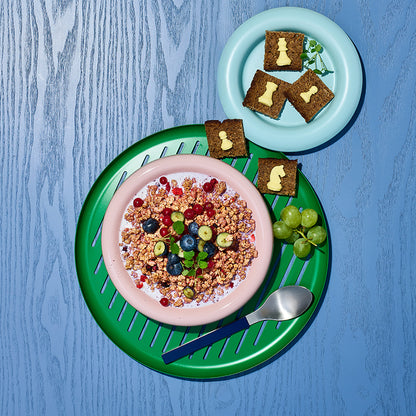 Blick von oben auf eine rosafarbene Müslischüssel mit Krunchy Waldbeere weniger Zucker von Bio-Knuspermüsli Hersteller Barnhouse, Johannisbeeren und Heidelbeeren. Die Schüssel steht auf einem grün-blauen Teller, auf dem auch noch ein blauer Löffel, grüne Weintrauben sowie kleine Vollkornbrot-Stücke mit Butter in Schachfiguren-Form liegen. Darüber liegt ein kleinerer, hellblauer Teller, auf dem weitere Vollkornbrot-Stücke mit Butter in Schachfiguren-Form liegen. Der Untergrund ist ein blauer Holztisch.