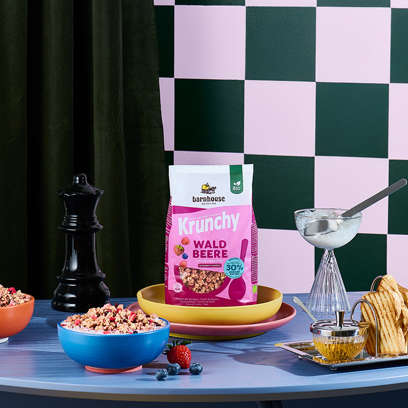Rosa-beerenfarbener Beutel Krunchy Waldbeere weniger Zucker von Barnhouse in gelber Schüssel auf rosa Teller.
Vorne eine blaue Schüssel mit Milch und dem besagten Bio-Knuspermüsli. Daneben eine Erdbeere und Heidelbeeren. Außerdem stehen noch ein Zuckerglas mit silbernem Löffel, ein silbernes Tablett mit Honig und Toastbrot sowie eine schwarze Pfeffermühle in Form einer Schach-Dame auf dem blauen Holztisch. Im Hintergrund eine Wand mit dunkelgrün-gräulichem Schachmuster sowie einem dunkelgrünen Vorhang.