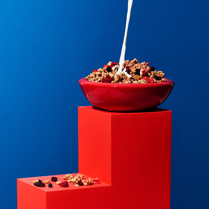 Roter Holzklotz und spiegelverehrter L-Form vor blauem Hintergrund. Auf der unteren "Stufe" liegen Bio-Knuspermüsli Cluster sowie Himbeeren, Heidelbeeren und Erdbeeren. Oben steht eine rote Schüssel mit Krunchy Waldbeere weniger Zucker mit Himbeeren, Heidelbeeren und Erdbeeren. Von oben fließt ein Milchstrahl in die Schüssel