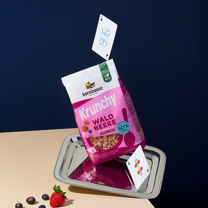 Silbernes Tablett, auf dem ein rosa-beerenfarbener Beutel Krunchy Waldbeere weniger Zucker von Bio-Knuspermüsli Hersteller Barnhouse steht. Darunter und darüber jeweils eine Spielkarte. Vorne links liegen Heidelbeeren und eine Erdbeere. Das Tablett liegt auf einem cremefarbenen Tisch, der Hintergrund ist dunkelblau.