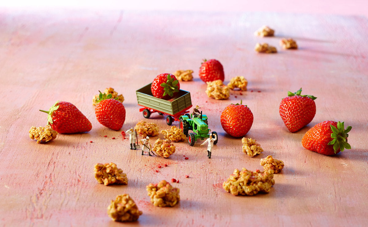 Miniaturbild, bei dem Erdbeeren und Knuspermüsli-Cluster auf einem leicht rosa-farbenen Holztisch liegen. 3 Männchen arbeiten mit Spaten auf dem "Feld". In der Mitte steht ein grüner Traktor mit Fahrer-Männchen, der eine frische Erdbeere mit einem grün-roten Anhänger fährt.