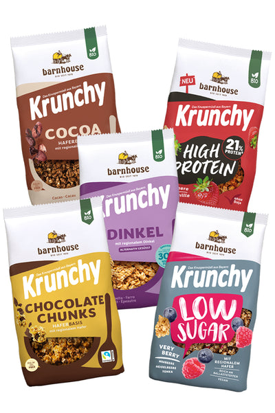 Blick von vorne auf die fünf Packungen Krunchy Cocoa weniger Zucker (braun-beige) High Protein (rot-schwarz), Dinkel weniger Zucker (violett), Chocolate Chunks (gelb-braun) und Low Sugar Very Berry (rot-blau)