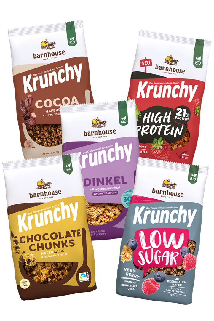 Blick von vorne auf die fünf Packungen Krunchy Cocoa weniger Zucker (braun-beige) High Protein (rot-schwarz), Dinkel weniger Zucker (violett), Chocolate Chunks (gelb-braun) und Low Sugar Very Berry (rot-blau)