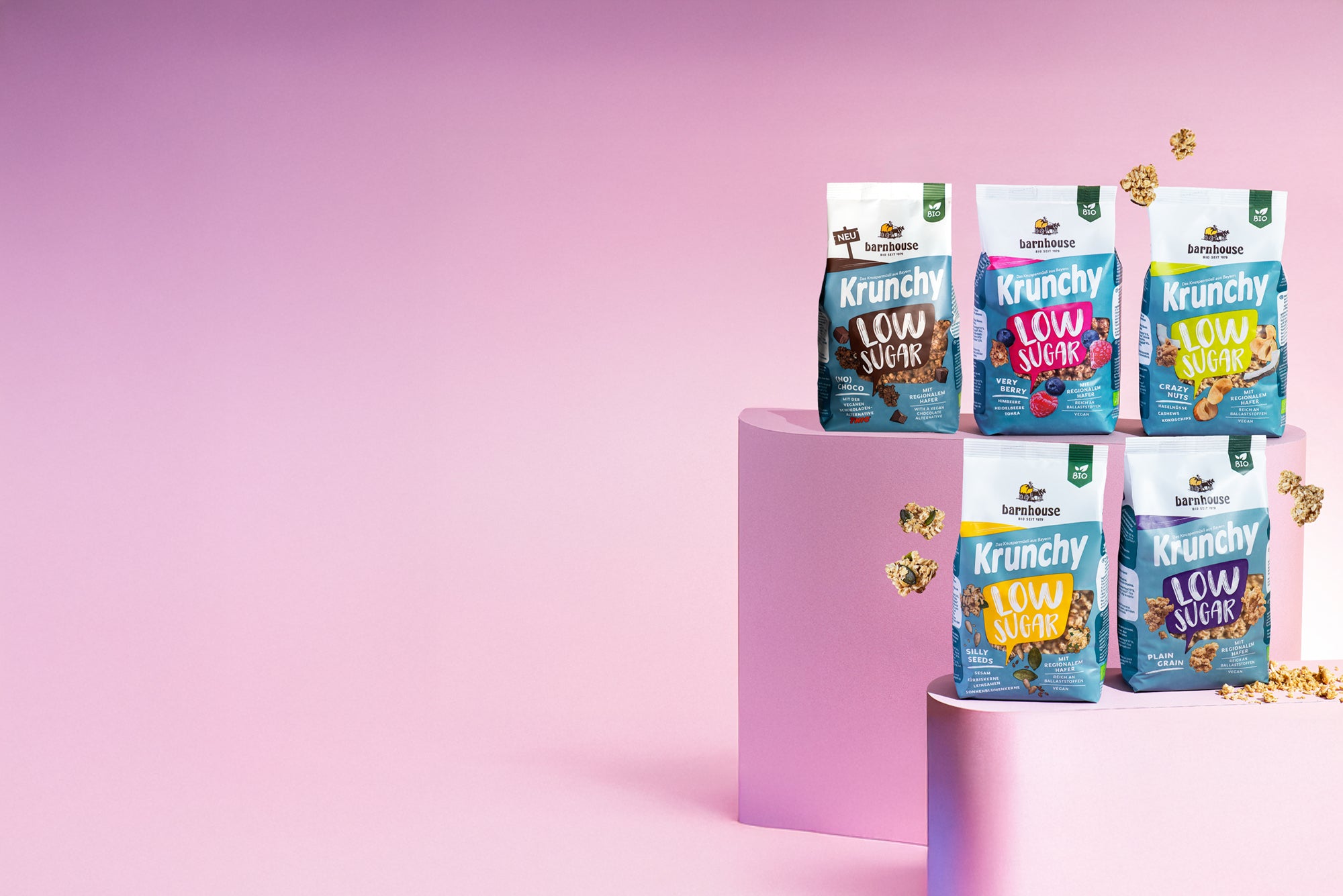 Blick von vorne auf die schräg angeordneten Packshots der fünf Low Sugar Krunchys Plain Grain, Crazy Nuts, Very Berry (No) Choco und Silly Seeds von Bio-Knuspermüsli-Hersteller Barnhouse, die auf zwei verschiedenen rosa Sockeln stehen. Außenrum fliegen Cluster.