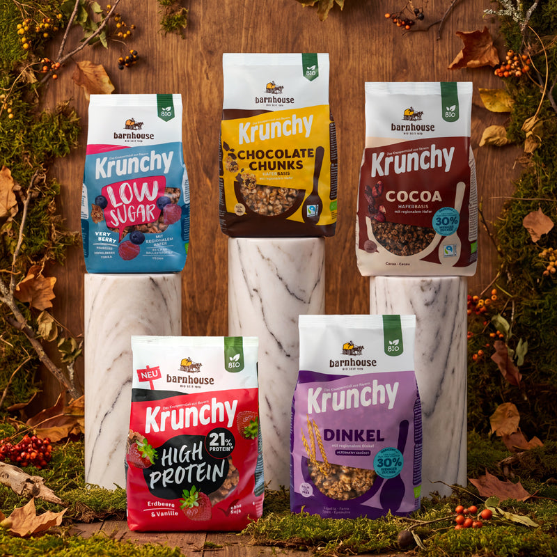 Blick von vorne auf die Packungen von Krunchy Low Sugar Very Berry (rot-blau), Chocolate Chunks (gelb-braun) und Cocoa weniger Zucker (braun-beige), die jeweils auf einer weißen Marmorsäule stehen. Davor stehen die Beutel von High Protein (rot-schwarz) und Dinkel weniger Zucker (violett) am Boden. Im Hintergrund eine Holzwand, die mit Ästen, Zweigen, Moos, Blättern und Beeren herbstlich dekoriert ist.