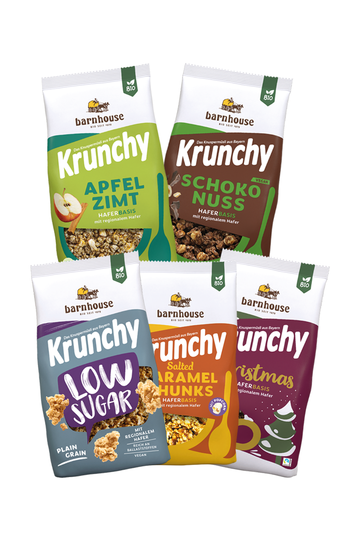 5 Krunchy-Beutel in Fächerform angerichtet. Oben überlappen sich Krunchy Apfel-Zimt und Schoko-Nuss. Darunter einer 3er Fächer mit Krunchy Low Sugar Plain Grain, Salted Caramel Chunks und Christmas.