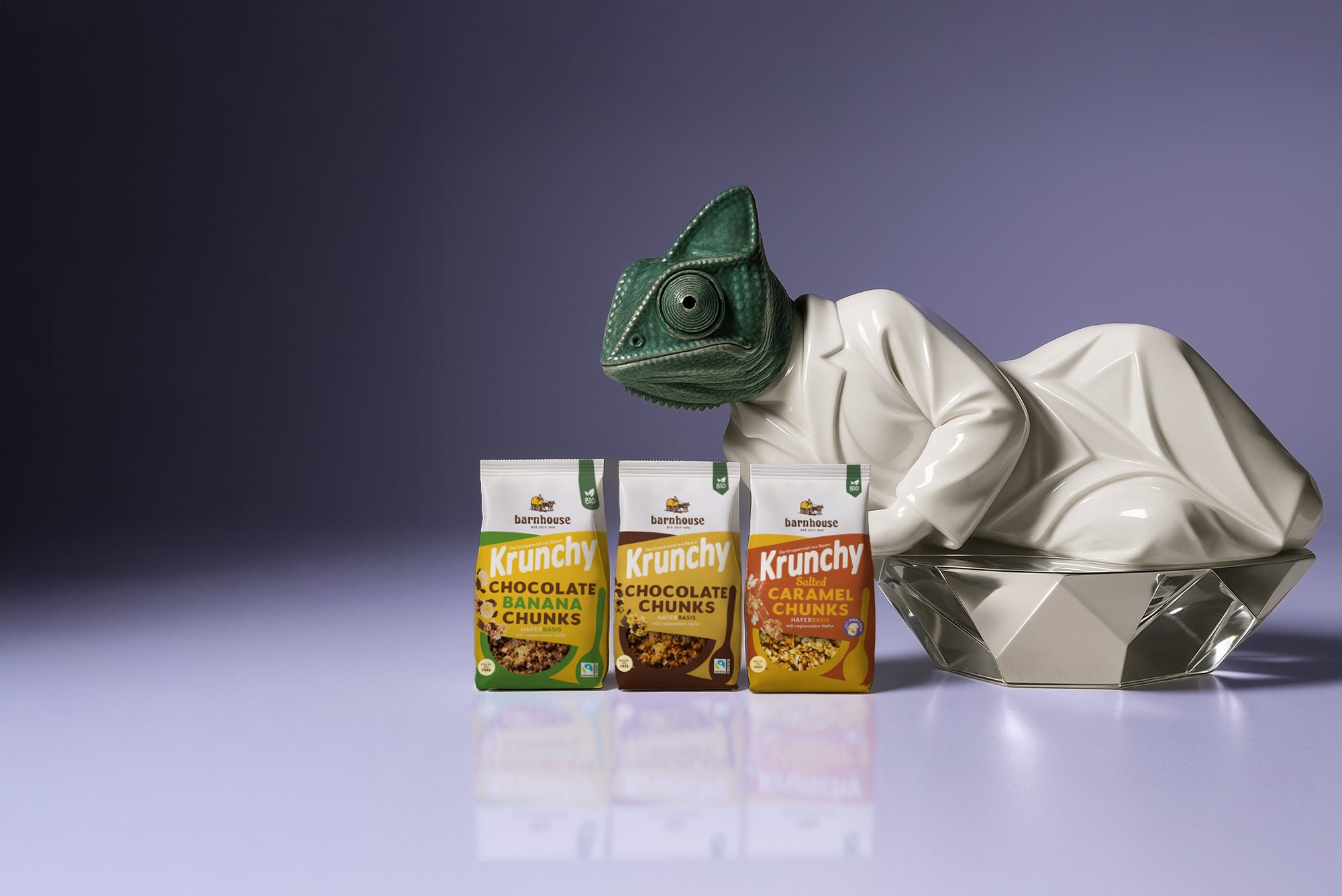 Chameleon-Skulptur in einem weißen Mantel auf einem silbernen Sockel. Daneben stehen die 3 Beutel von Krunchy Chocolate Banana Chunks, Chocolate Chunks und Salted Caramel Chunks.