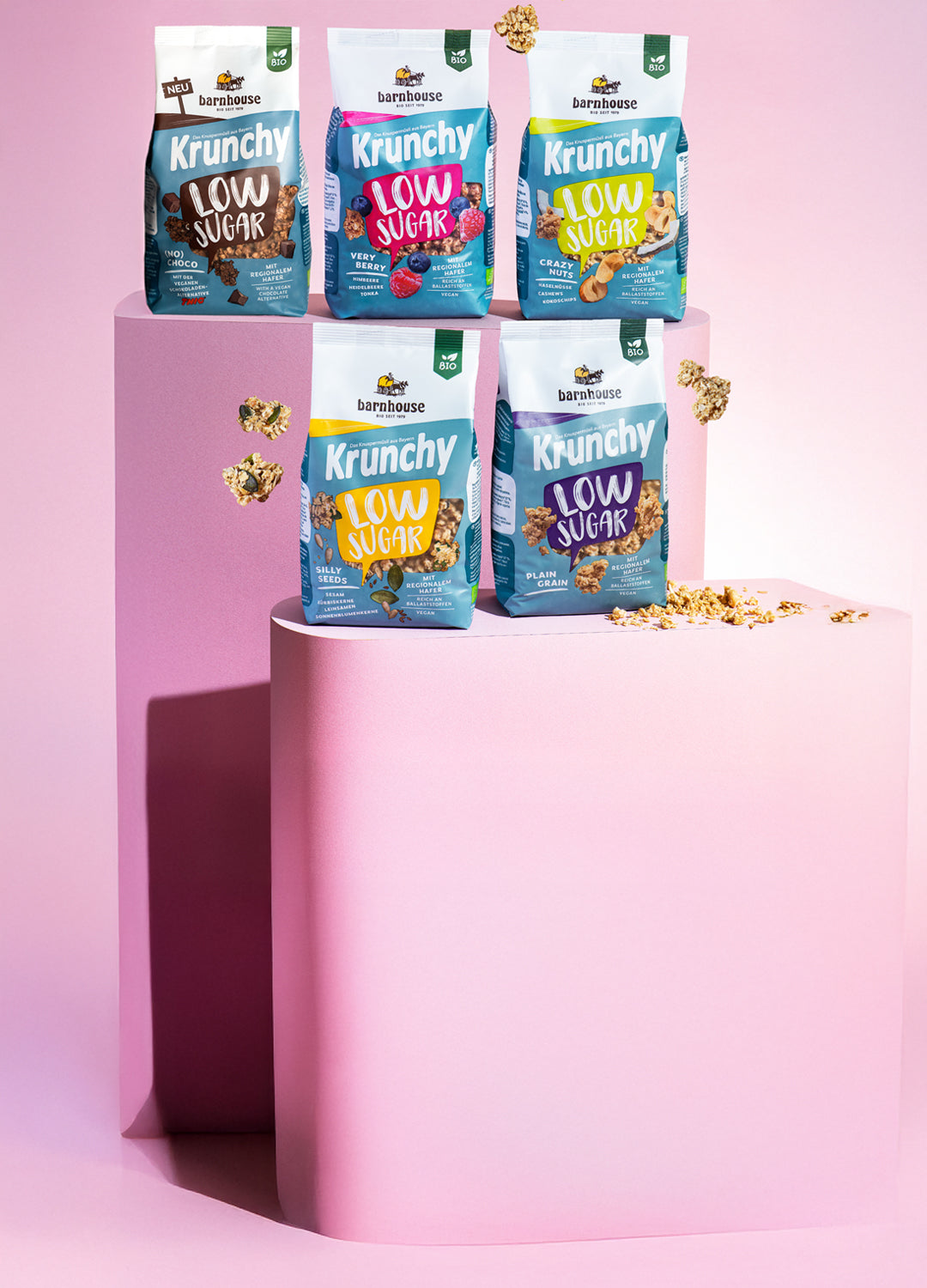 Blick von vorne auf die schräg angeordneten Packshots der fünf Low Sugar Krunchys Plain Grain, Crazy Nuts, Very Berry (No) Choco und Silly Seeds von Bio-Knuspermüsli-Hersteller Barnhouse, die auf zwei verschiedenen rosa Sockeln stehen. Außenrum fliegen Cluster.