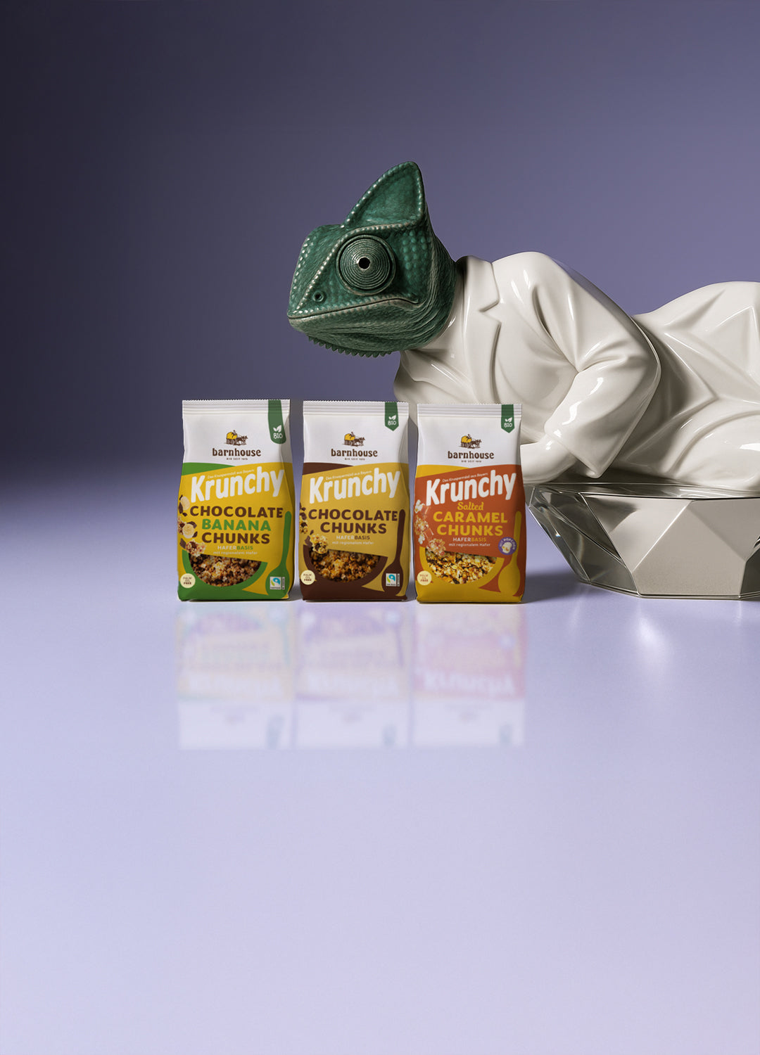 Chameleon-Skulptur in einem weißen Mantel auf einem silbernen Sockel. Daneben stehen die 3 Beutel von Krunchy Chocolate Banana Chunks, Chocolate Chunks und Salted Caramel Chunks.